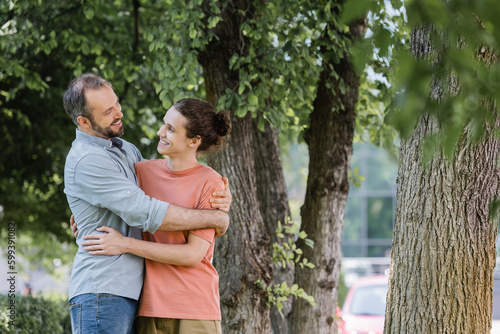 joyful father with beard hugging happy teenager son in green park.