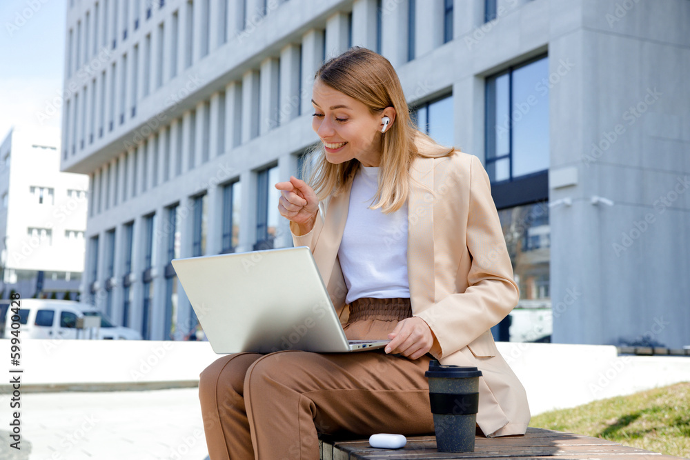 Smiling businesswoman using modern laptop for video call while sitting on bench outdoors. Lady worker in earphones making hello gesture and smiling during online communication Smiling businesswoman using modern laptop for video call while sitting on bench outdoors. Lady worker in earphones making hello gesture and smiling during online communication