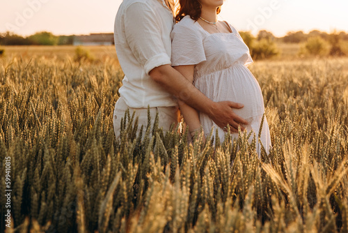 A guy hugs a pregnant girl's stomach in the middle of a wheat field at sunset in summer. golden hour Waiting for the birth of a child by a husband and wife. Support of husband
