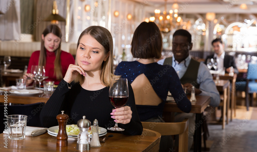 Pensive young woman sitting alone in restaurant table with glass of red wine