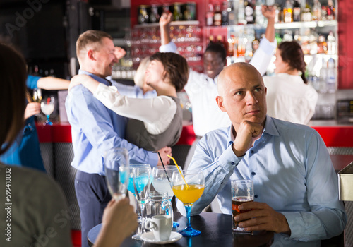 Troubled man sitting alone at office party in nightclub on background with cheerful workmates