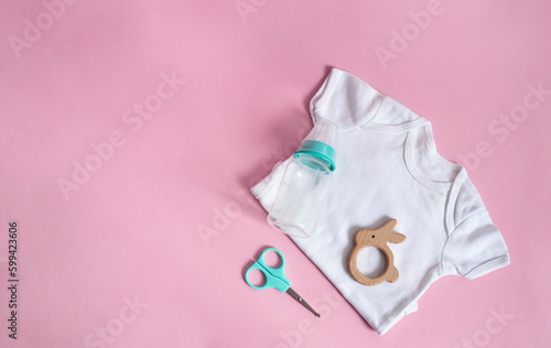 baby t-shirt with a bottle, a wooden teether and scissors for little ones on a pink background. Baby care, daily hygiene and care. Place for text. top view, flat lay