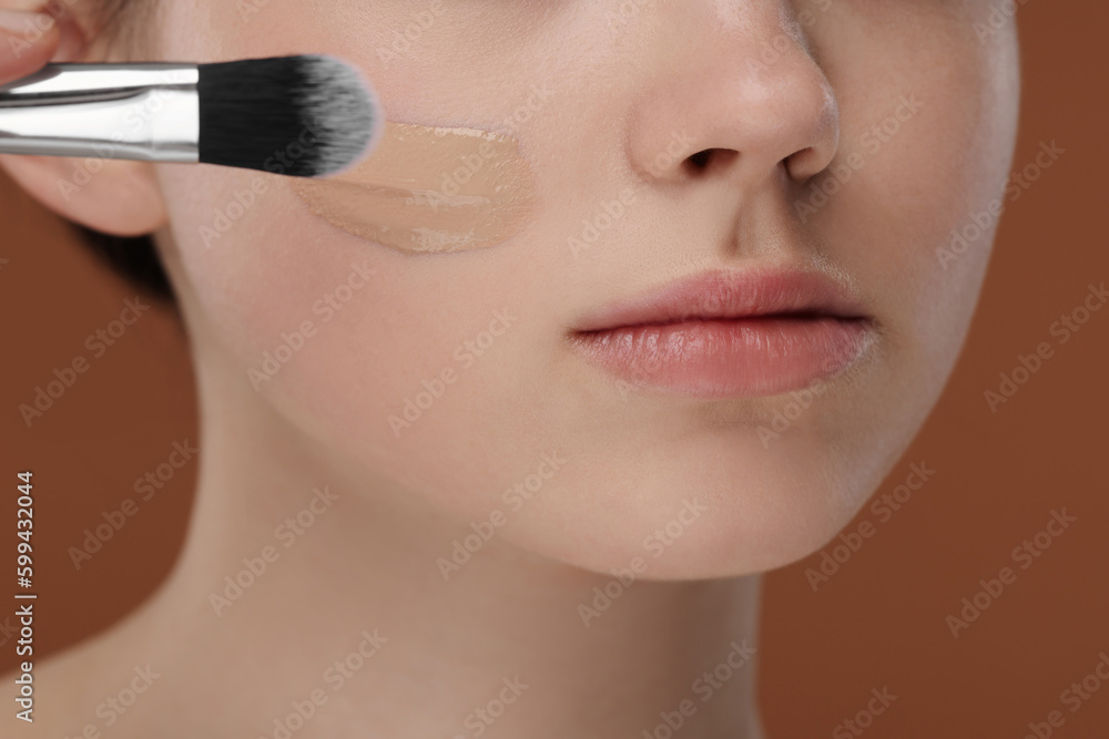 Teenage girl applying foundation on face with brush against brown background, closeup Teenage girl applying foundation on face with brush against brown background, closeup
