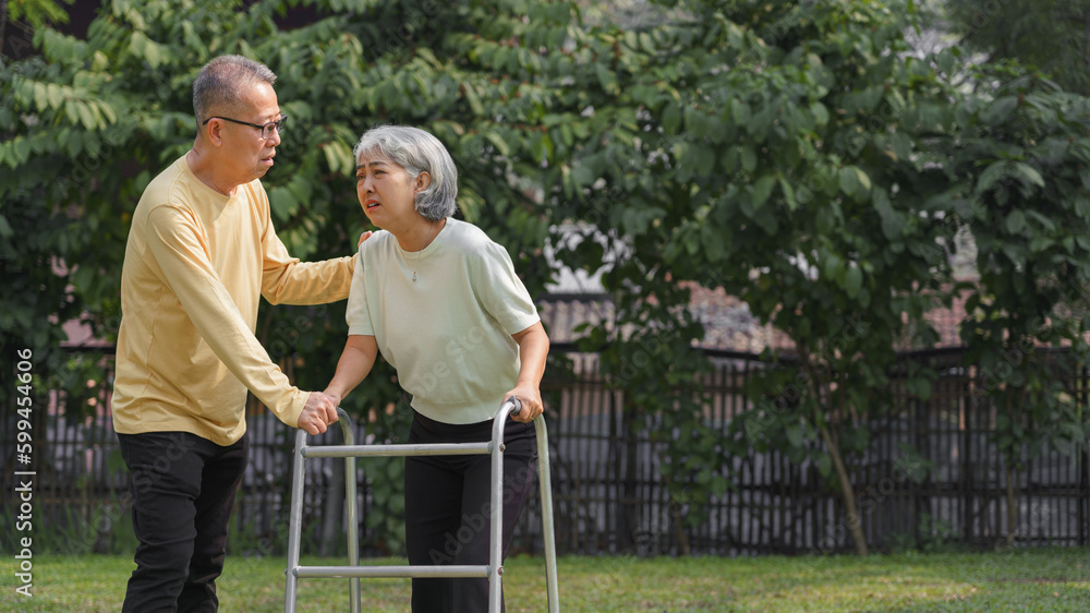Senior man helping beloved wife walking with walker frame while doing rehab physical in the park Senior man helping beloved wife walking with walker frame while doing rehab physical in the park