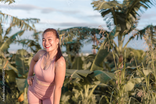 A fit woman wearing a workout outfit and posing by leaning to the right with one hand on hip and the other hand on the legs, Tall grass and coconut trees in the background © Mdv Edwards