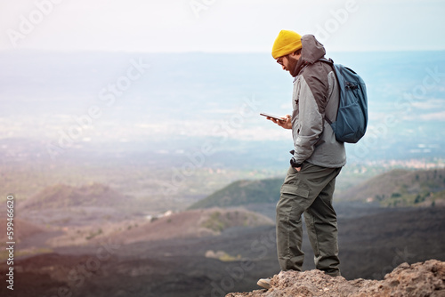 A man 30 y old is on a peak, at a high altitude,  with a beautiful view, but he is looking at the smartphone and not at the landscape.