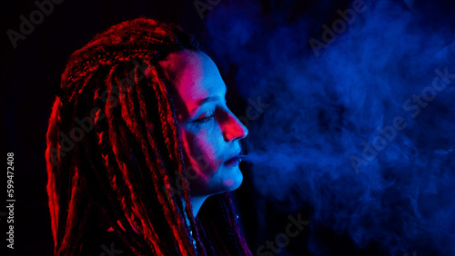Profile of caucasian girl with dreadlocks smokes a vape in red blue light. 