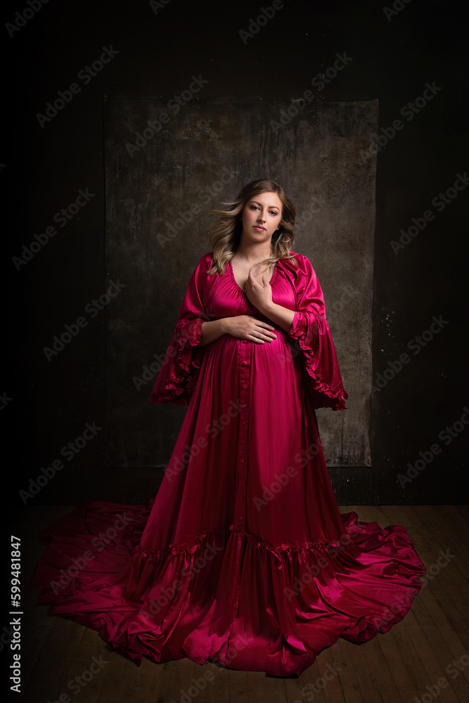 Beautiful pregnant woman with long hair wind blown hair wearing long flowing maternity dress with low key dramatic studio lighting and neutral expression in delicate pose