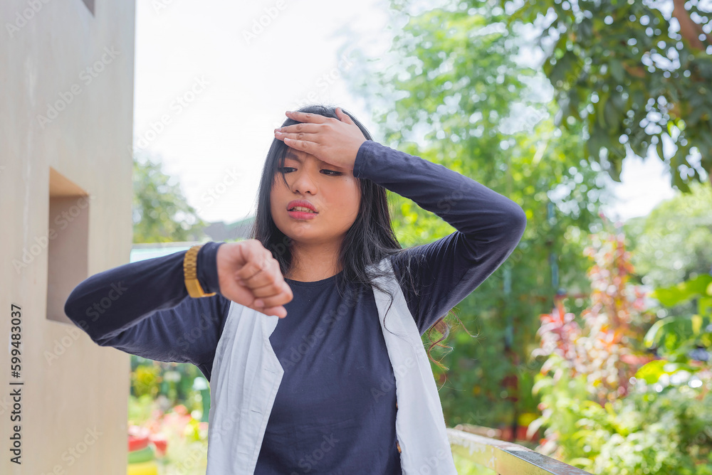 Naklejka premium A close-up of a lovely lady in a cardigan and navy blue long-sleeved shirt, facing the camera with head up, palm on her forehead while checking her wrist watch.