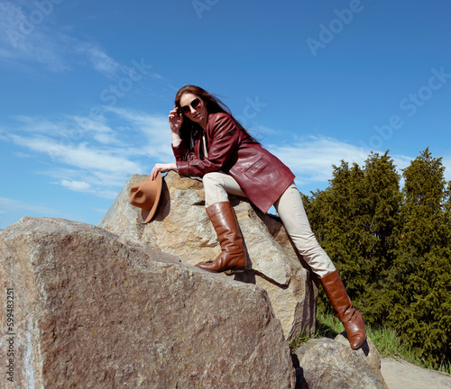 A lonely adult woman stands on the top of a cliff, looking into the vast expanses of the azure sky. A woman with long hair in a leather jacket stands on the rocks near the forest