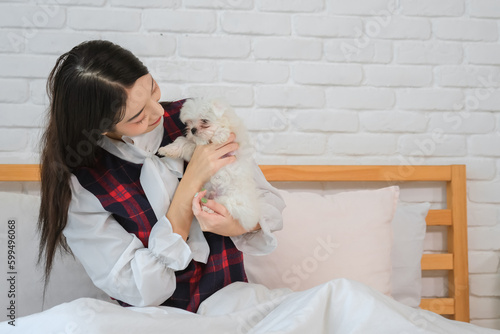 Attractive young asian woman holding white dog on bed in bedroom affectionately.
