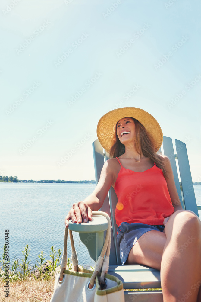 Fototapeta premium Nothing and nobody can spoil her mood. a cheerful young woman wearing a hat while being seated on a chair next to a lake outside in the sun.