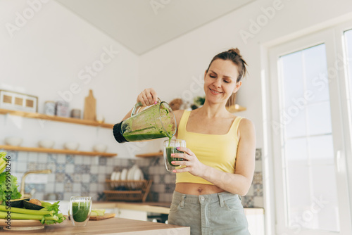 Happy woman on detox diet, pouring green cocktail from mixer into glass in kitchen