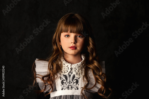 Portrait of cover girl in doll image 9-10 year old on black background, frozen position looking at camera. Adorable kid with curly long hair isolated in shadow. Theatrical concept. Copy ad text space