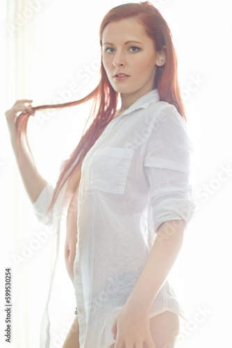 Shes a firery beauty. Portrait of an attractive redheaded woman wearing an unbuttoned shirt standing in a bedroom looking sexy while standing in her underwear. Young female posing semi nude. photo