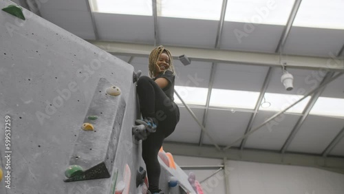 Smiling Woman Reaching Top Of Climbing Wall In Indoor Activity Centre Watched By Coach