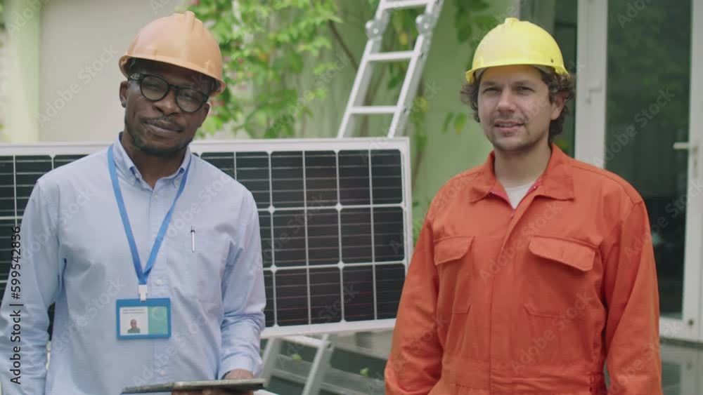 Medium shot of cheerful African American engineer and young Caucasian worker posing together for camera with smile by solar panel outdoors Medium shot of cheerful African American engineer and young Caucasian worker posing together for camera with smile by solar panel outdoors