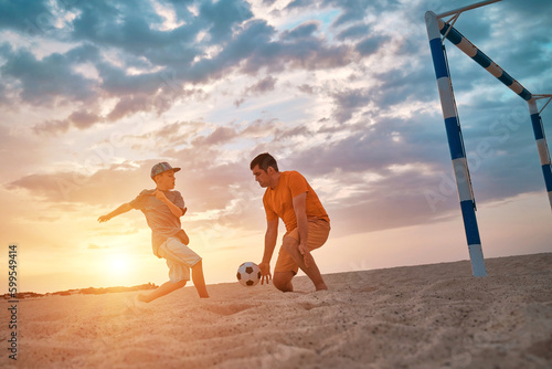 Father and Son playing football, family fun outdoors players in soccer in dynamic action have fun playing football in the beach, summer day under sunlight. 