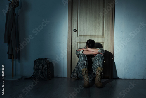 Canvas Print Lonely soldier in uniform sitting near backpack and door at home at night