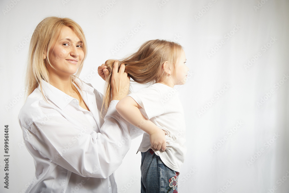 Close portrait of blonde mother and daughter where mom braids hair and makes ponytail and hairstyle for her girl on white background in studio. The concept of love, friendship, caring in the family Close portrait of blonde mother and daughter where mom braids hair and makes ponytail and hairstyle for her girl on white background in studio. The concept of love, friendship, caring in the family