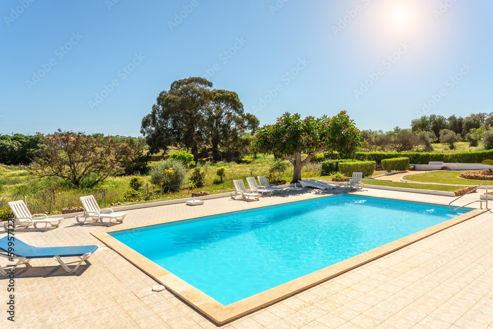 Private swimming pool with sun loungers and garden around. Sunny day.
