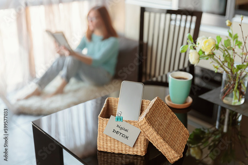 Smartphone is in separate wicker box with inscription digital detox on table. Woman reading book in background. Stop using digital gadgets. Mental and digital detox concept photo