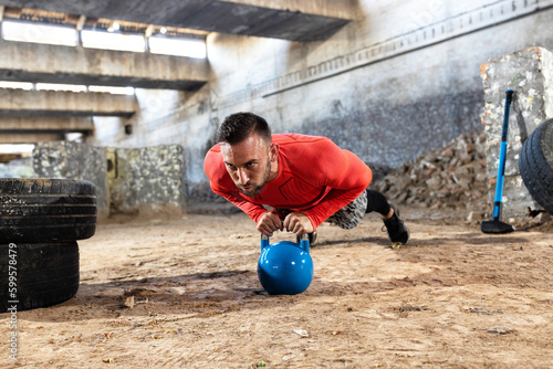 Handsome young man doing kettlebell pushups, additionally building up abdominal, shoulder and chest strength, closed industrial complex factory training