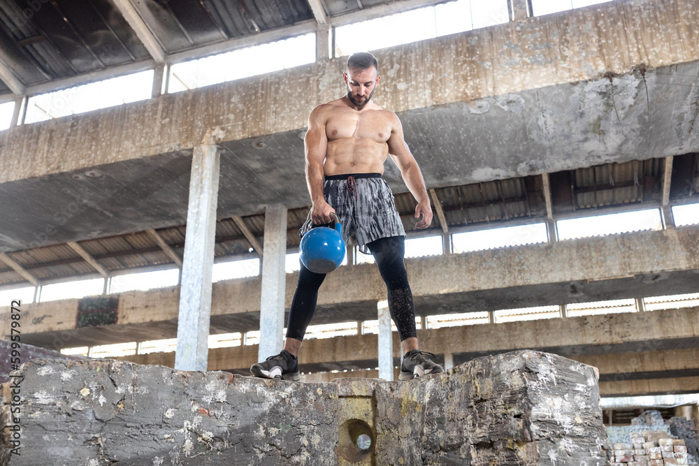 Strong ripped guy standing on big concrete blocks in a old factory preparing to do kettlebell single arm dead lift