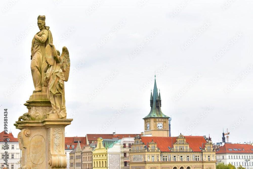 Ancient catholic sculptures on a pedestal and old buildings with red roofs and tower with the green top and clock. Statues on biblical subjects. Charles Bridge. Prague, Czech Republic, October 2022.