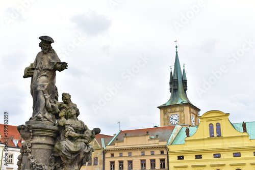 Ancient catholic sculptures on a pedestal and old buildings with green roofs and tower with the green top and clock. Statues on biblical subjects. Charles Bridge. Prague, Czech Republic, October 2022.