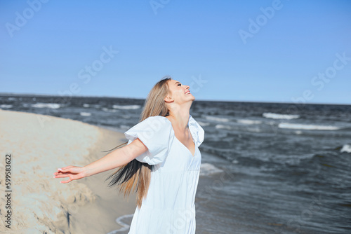 Happy blonde woman is on the ocean beach in a white dress and sunglasses  raising hands