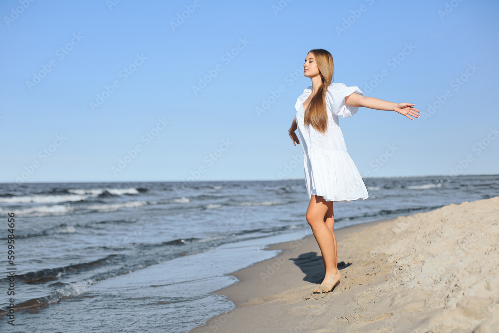 Happy blonde woman is on the ocean beach in a white dress and sunglasses, raising hands