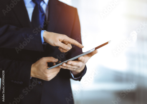 Business people use a tablet computer for discussion of their new project, standing in a sunny modern office. Unknown businessman or male entrepreneur with a colleague at workplace