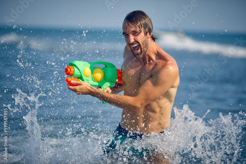 Young handsome man at beach, in water shooting with watergun making excited expression. Man on summer vacation. Holiday, fun, lifestyle concept.