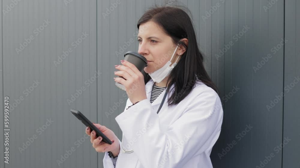 Portrait of smiling young female doctor in a medical mask and white coat with stethoscope who using smartphone and drinking coffee outdoors on backyard of hospital Portrait of smiling young female doctor in a medical mask and white coat with stethoscope who using smartphone and drinking coffee outdoors on backyard of hospital