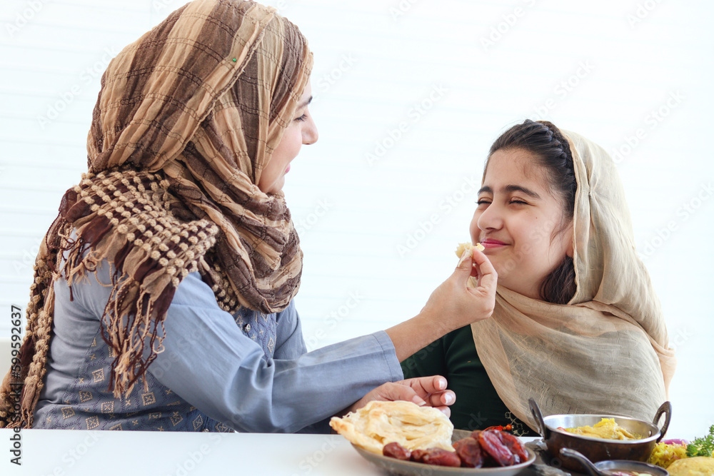 Adorable Muslim happy girl sits at kitchen table, kid with hijab enjoy eating traditional Islamic halal food with mother hand on white background, mom feeding child daughter, warm love in family.