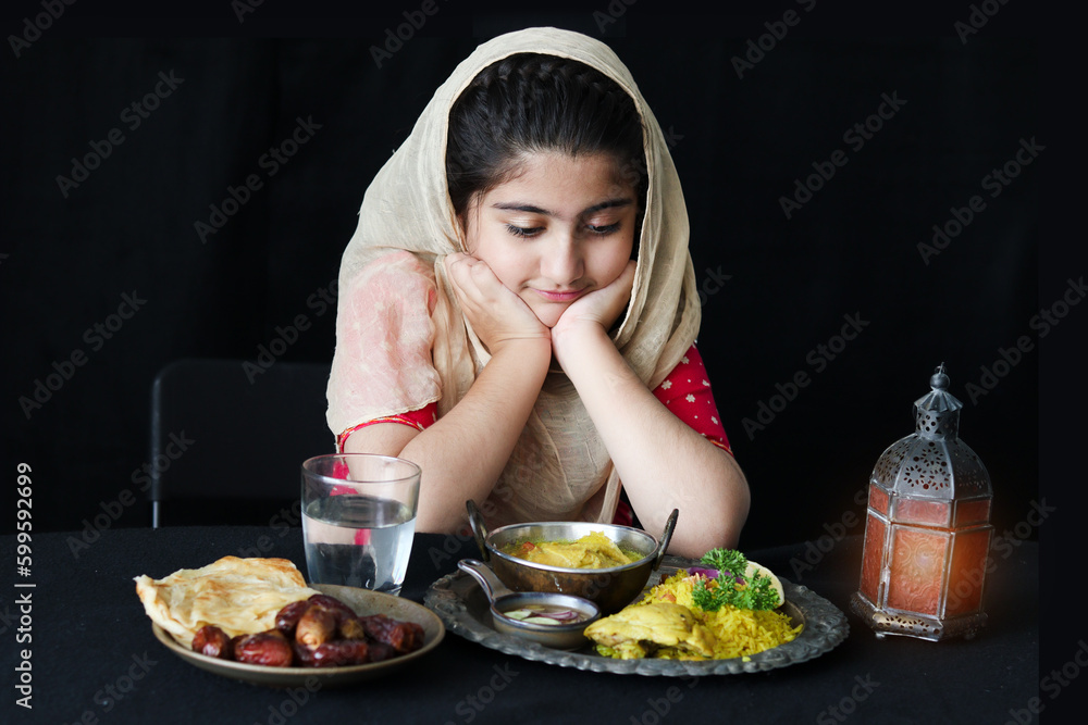 Adorable happy Muslim girl with beautiful eyes wearing hijab and traditional costume resting chin in hands at kitchen table, portrait of kid eating Islamic halal food meal on dark black background. Adorable happy Muslim girl with beautiful eyes wearing hijab and traditional costume resting chin in hands at kitchen table, portrait of kid eating Islamic halal food meal on dark black background.