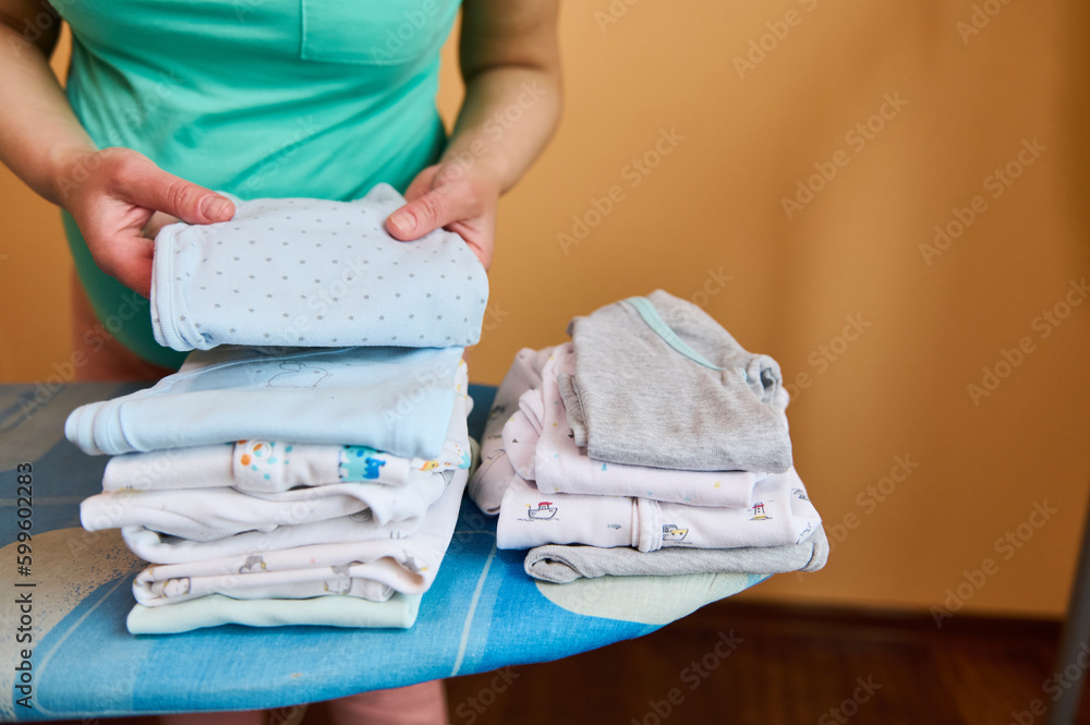 Closeup of expectant mother, loving caring pregnant woman in home wear,  standing by ironing board, arranging clean ironed clothes for her newborn  baby. Happy moments of carefree pregnancy. Maternity Stock Photo