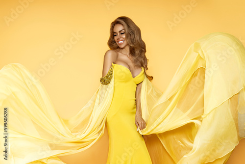 Fashion Woman in Yellow fluttering Dress. Glamour Model is posing in Long Silk Fabric flying on Wind. Elegance.