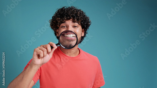 funny latin or arabian man shows white teeth and smiles through a magnifying glass in coral t-shirt on blue studio background