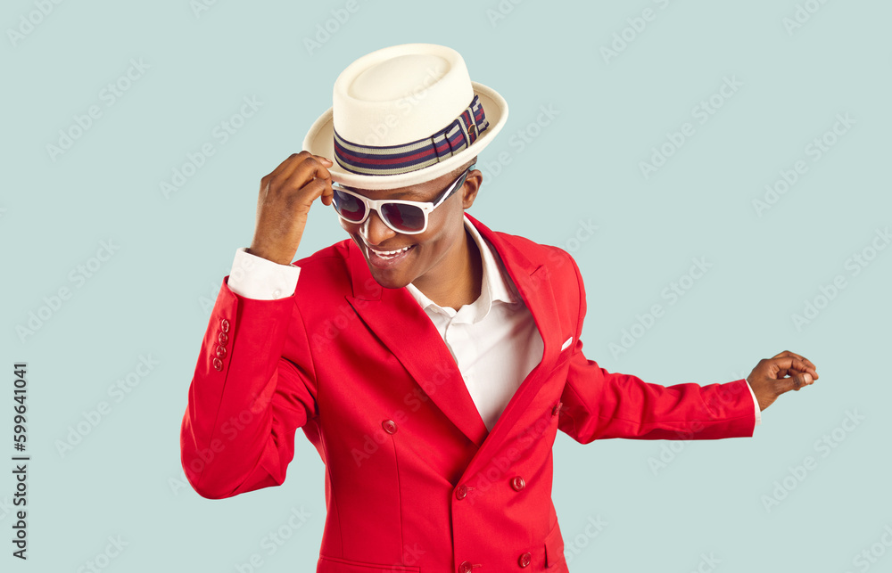 Time for party. Happy refined and stylish african man having fun dancing on pastel light blue background. Joyful positive man in red suit, white shirt, sunglasses holding on to hat having fun at party Time for party. Happy refined and stylish african man having fun dancing on pastel light blue background. Joyful positive man in red suit, white shirt, sunglasses holding on to hat having fun at party