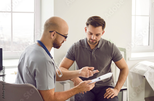 Doctor talks to his patient after a medical examination at the clinic. Young male physician shows a clipboard with the results of some medical tests they did and explains what this all means photo