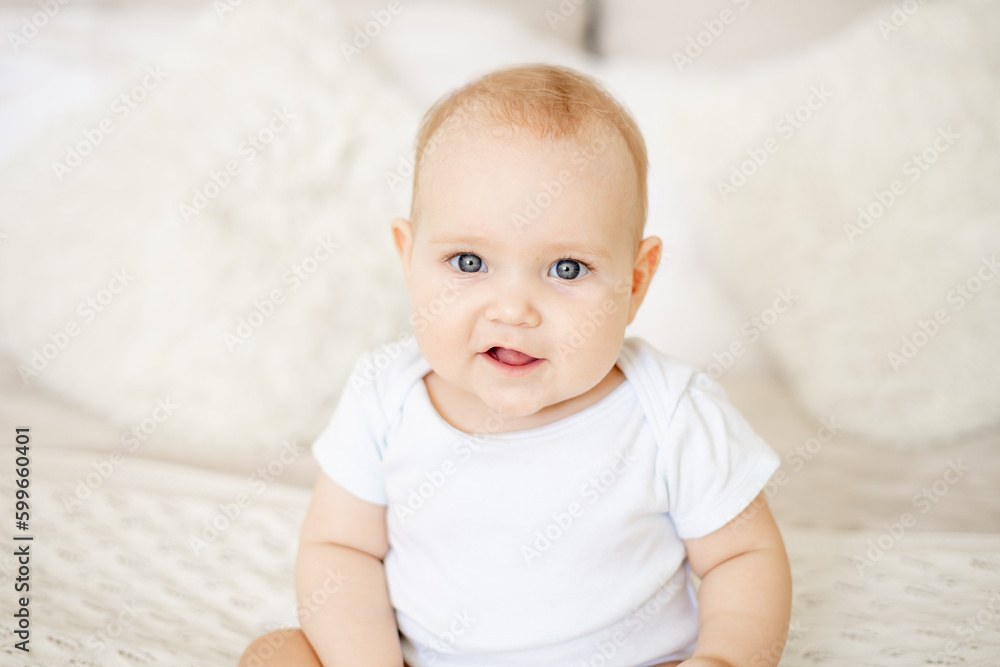 portrait close-up of a baby boy or girl of six months sitting at home on a bed in a bright bedroom and smiling or laughing, a happy newborn in a white bodysuit with blue eyes