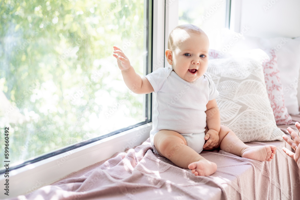 portrait of a close-up baby boy or girl of six months sitting at the window of the house in a bright room and smiling, a happy newborn in a white bodysuit at the window portrait of a close-up baby boy or girl of six months sitting at the window of the house in a bright room and smiling, a happy newborn in a white bodysuit at the window