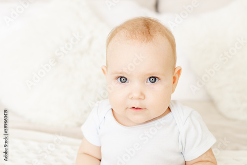 portrait close-up of a baby boy or girl of six months sitting at home on a bed in a bright bedroom and smiling or laughing, a happy newborn in a white bodysuit with blue eyes