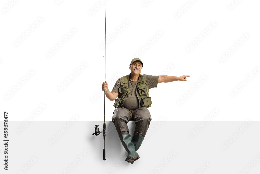 Cheerful mature fisherman with a fishing rod sitting on a blank panel and pointing to the side Cheerful mature fisherman with a fishing rod sitting on a blank panel and pointing to the side