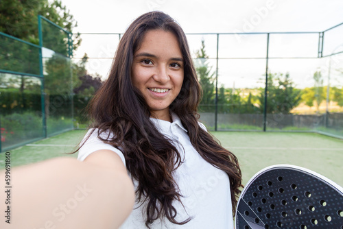 Young smiling woman taking a selfie with smart phone on padel court with racket