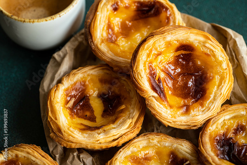 Pile of fresh traditional Portuguese pastel de nata on paper bag and cup of espresso on green surface.