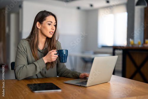 Young beautiful businesswoman using laptop computer and holding cup of coffee. Photo of female working on laptop device or reading news while drinking coffee in home office.