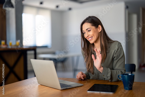 Young businesswoman employee, female HR manager waving hand looking at laptop during virtual video conference call sitting at desk in home office.